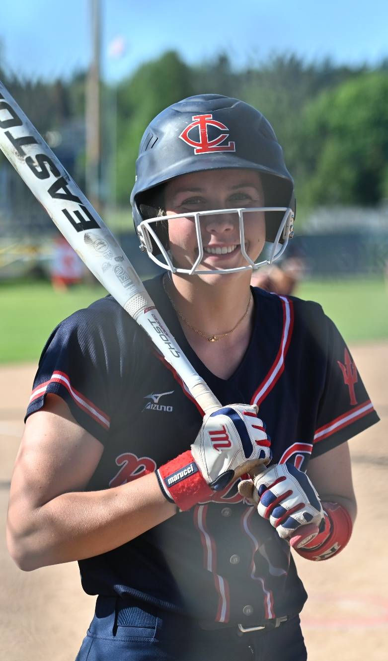 LCC female softball player wearing a blue LCC batters helmet and holding a white baseball bat over her right shoulder, wearing an LCC softball blue and red jersey and white baseball gloves; smiling for a photograph. 