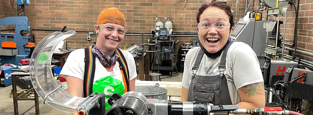 A photo of two students in the mech lab smiling for the camera. They are both wearing safety gear.