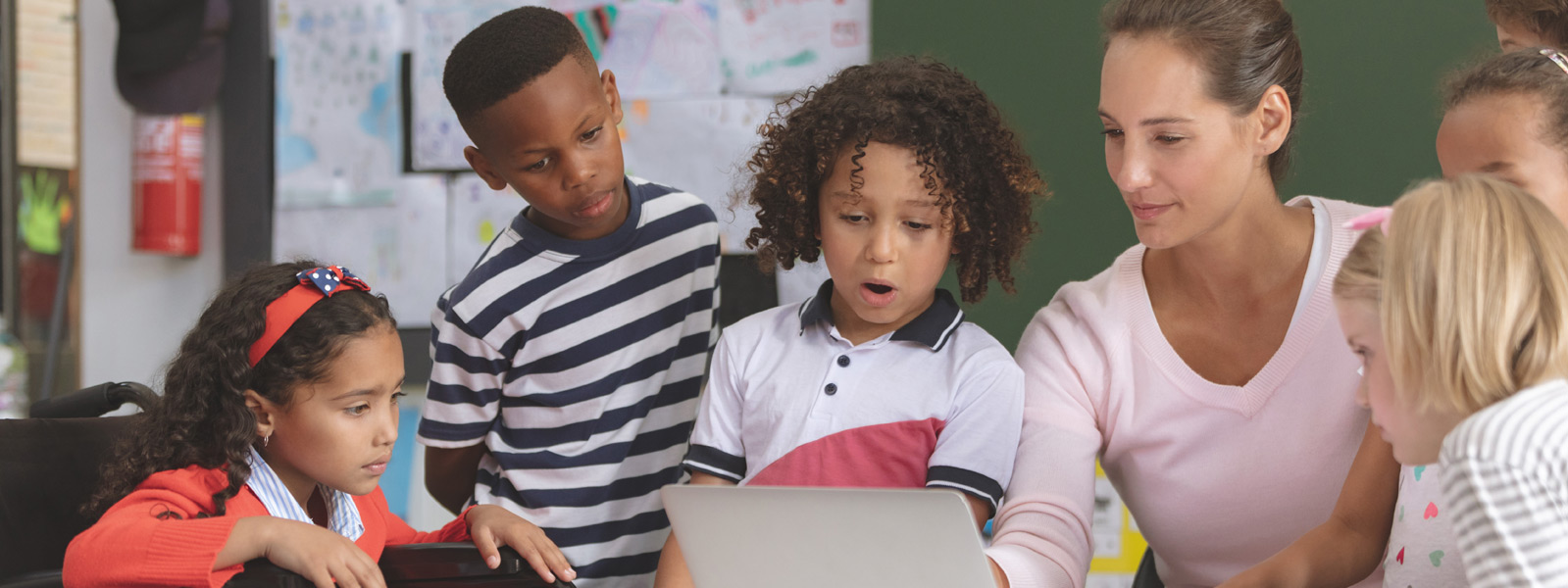 Female teacher at a desk with a laptop showing a group of students something on the laptop.