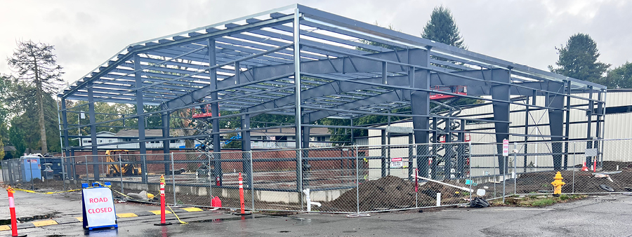 sports facility frame and structure under construction. Dark skies and rain water on the ground. Road closure sign and construction fencing around the constructed building.