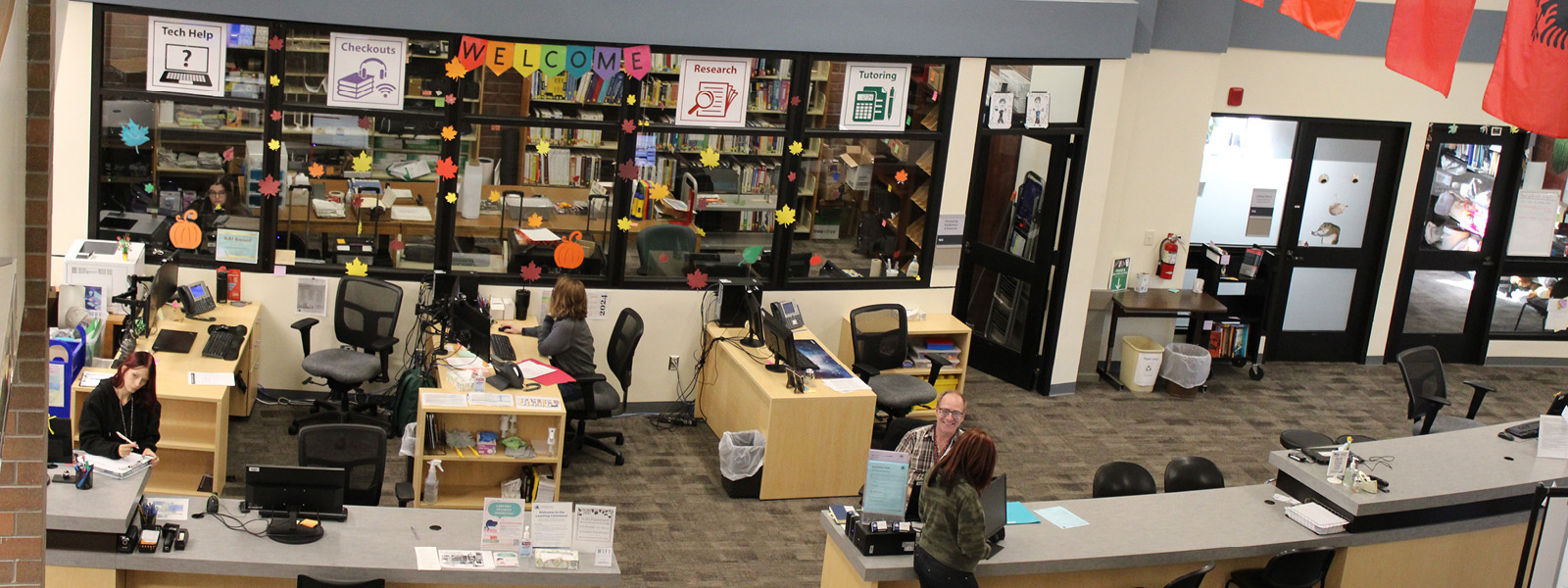 A welcoming library environment with workstations, decorated windows, and staff assisting visitors at the learning commons help desk.