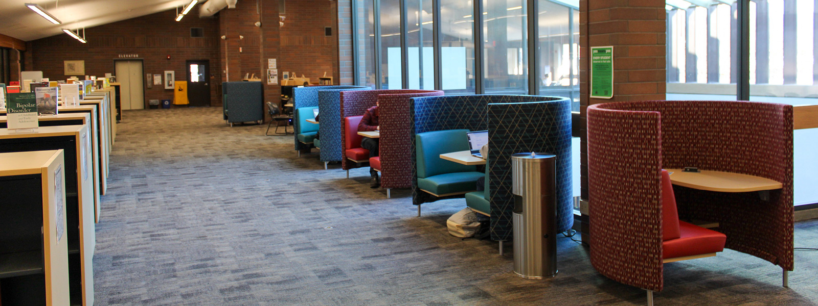 Spacious upstairs interior, of Alan Thompson Library featuring colorful study pods, a row of bookshelves, and large windows letting in natural light.