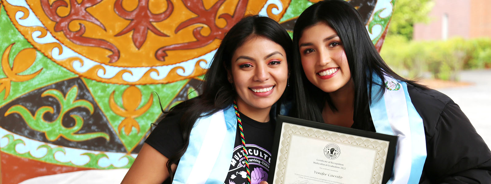 Two Latina Women standing in front of a colorful banner with Blue Stolls around their necks and holding Certificates for the Multicultural graduation recognition