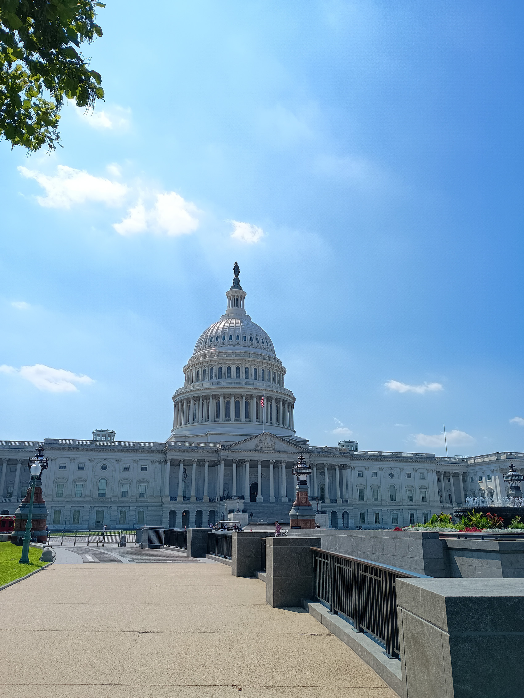 Yuna アメリカ議会図書館にて 薄い雲が浮かぶ鮮やかな青空の下、米国連邦議会議事堂の象徴的な白い新古典主義様式のファサードを、アイレベルのワイドショットで捉えています。