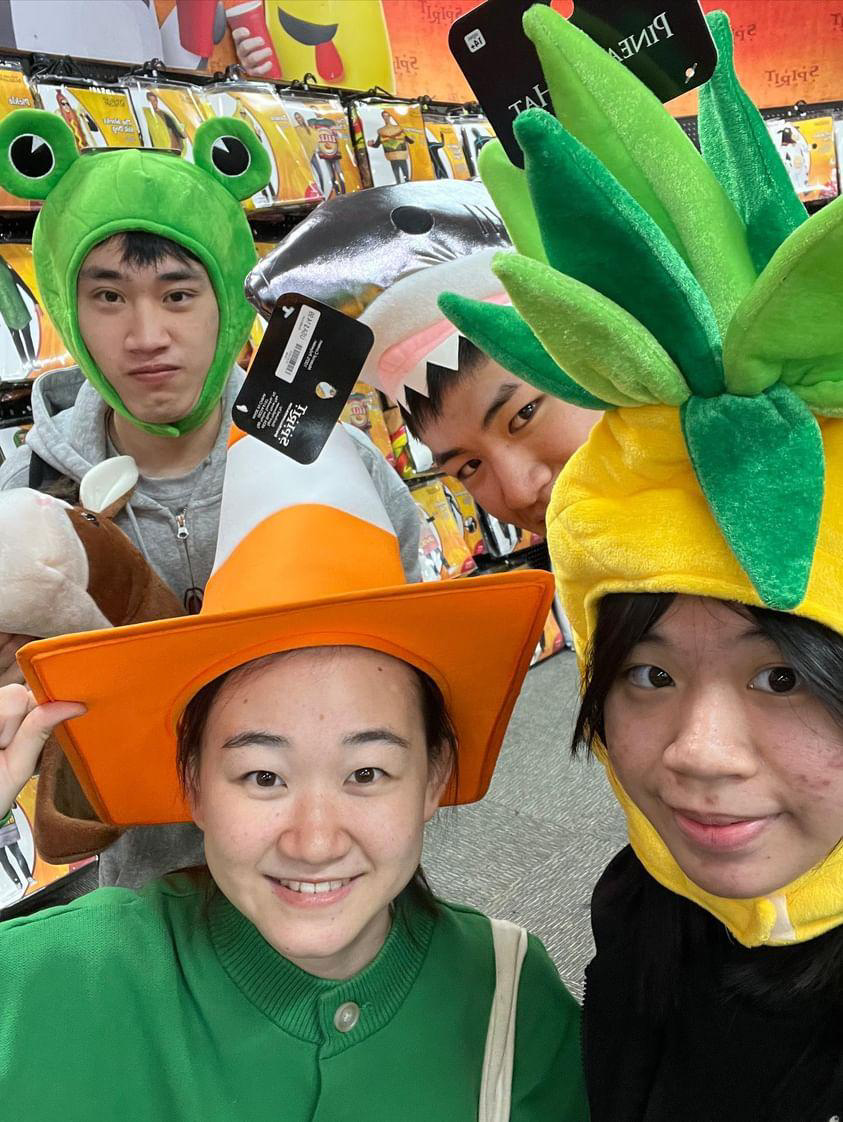 4 International student in spirit Halloween wearing funny hats, 1 boy is wearing a frog hat, 1 boy is wearing a shark hat, 1 girl is wearing a traffic cone hat, and 1 girl is wearing a pineapple hat