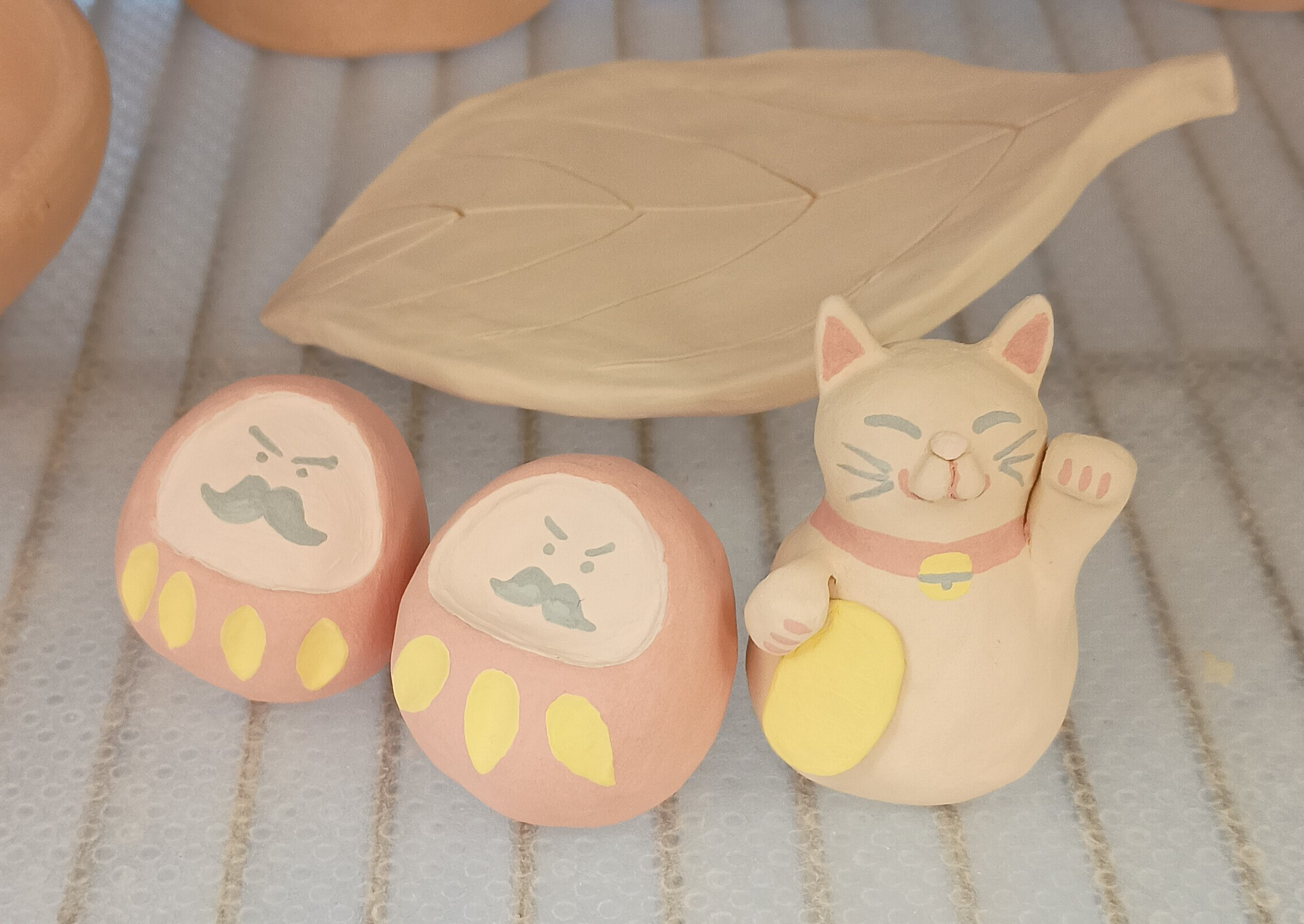 Ceramics A group of small, light peach-colored ceramic figurines, including two scowling Daruma dolls and a waving Maneki-neko lucky cat, sits in front of a leaf-shaped ceramic dish.