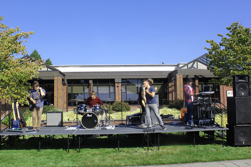 Live Music an outdoor musical performance featuring a band playing on a stage set up on a lawn in front of a modern single-story brick and glass building under a clear blue sky.