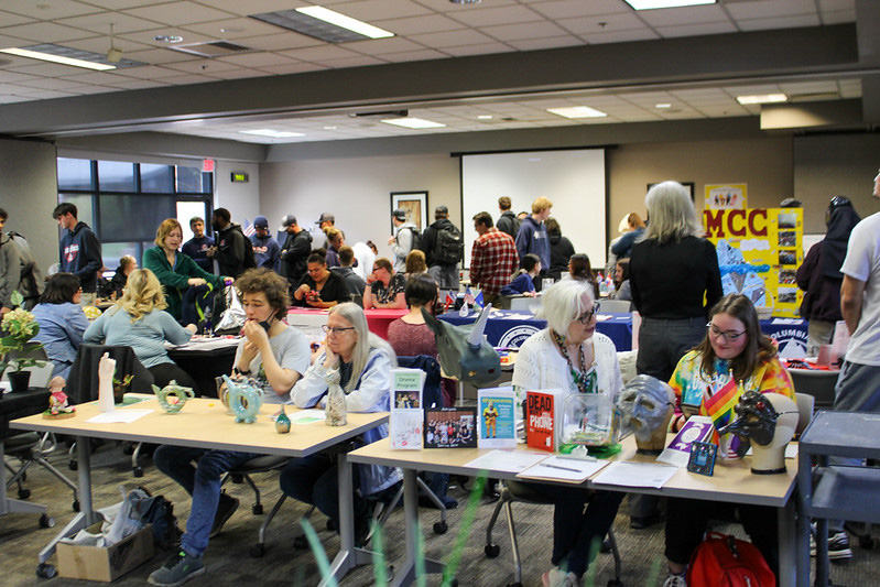 Club Fair LCC's club fair held in a large indoor room with tables displaying club information, promotional items, and student attendees interacting.