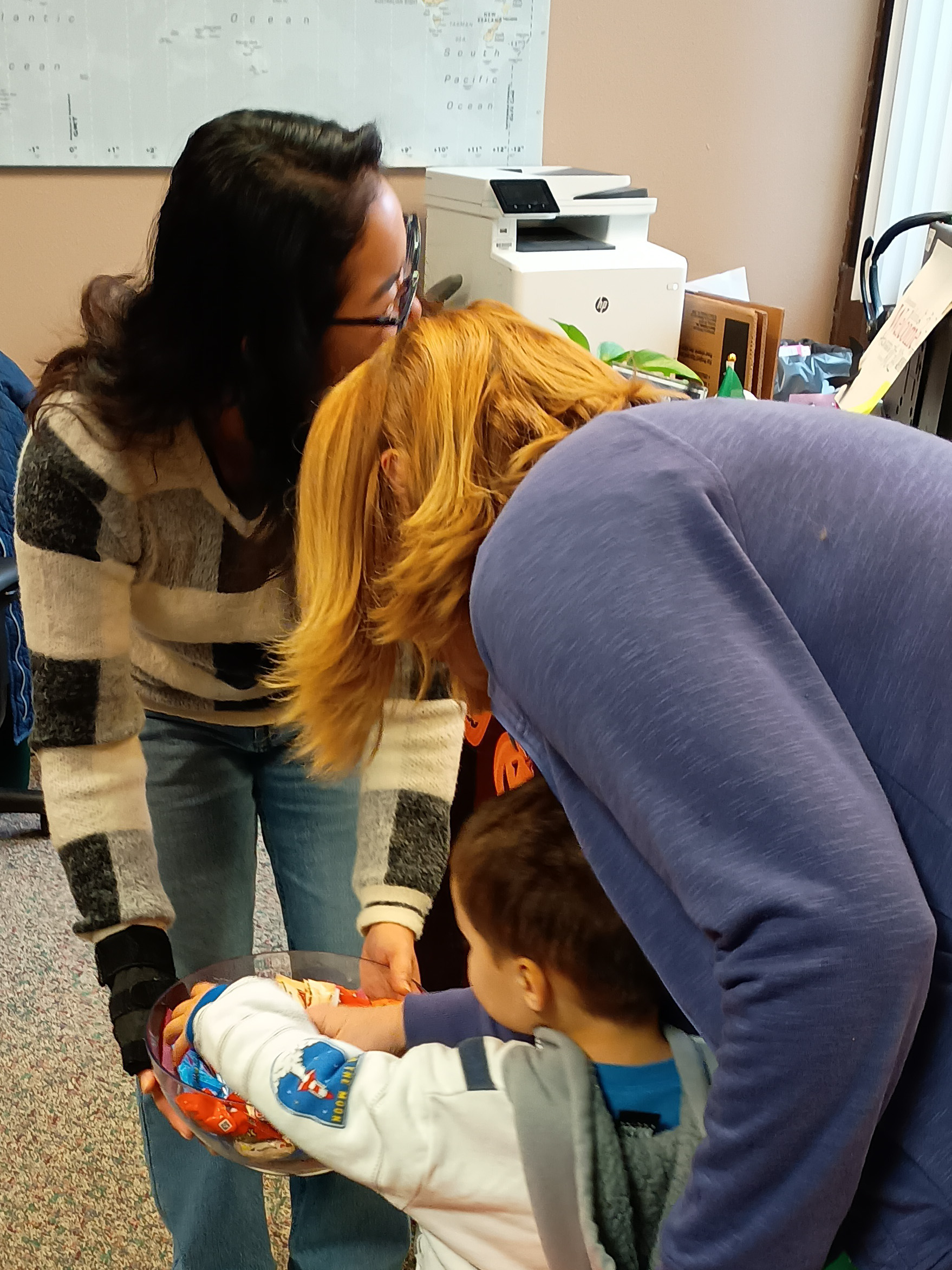 A Small child with and adult trick or treating at lcc, the small child is grabbing candy out of a candy bowl being held by a woman