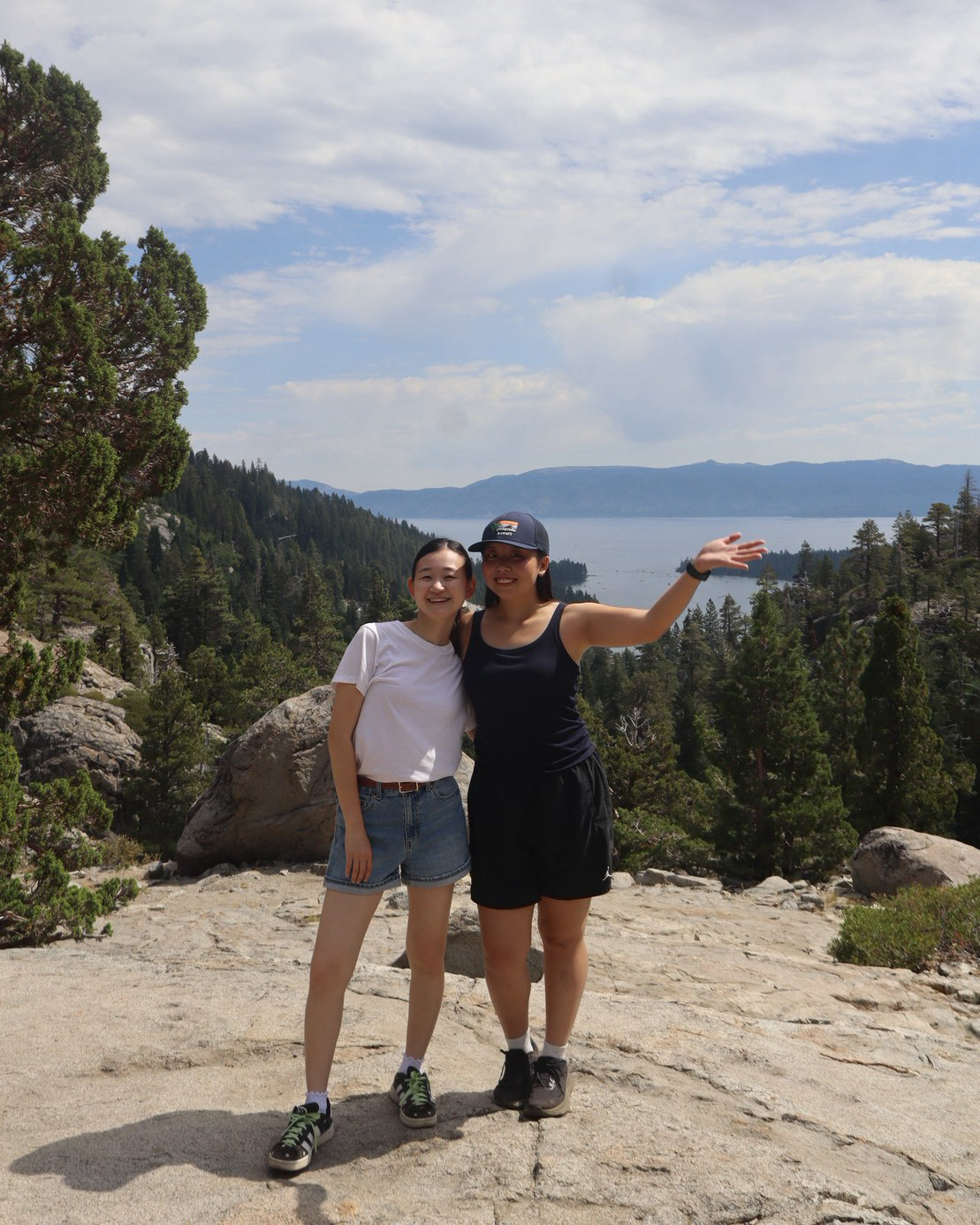 2 girls wearing t-shirts and shorts standing on a large flat rock, with a view out onto lake tahoe behind them