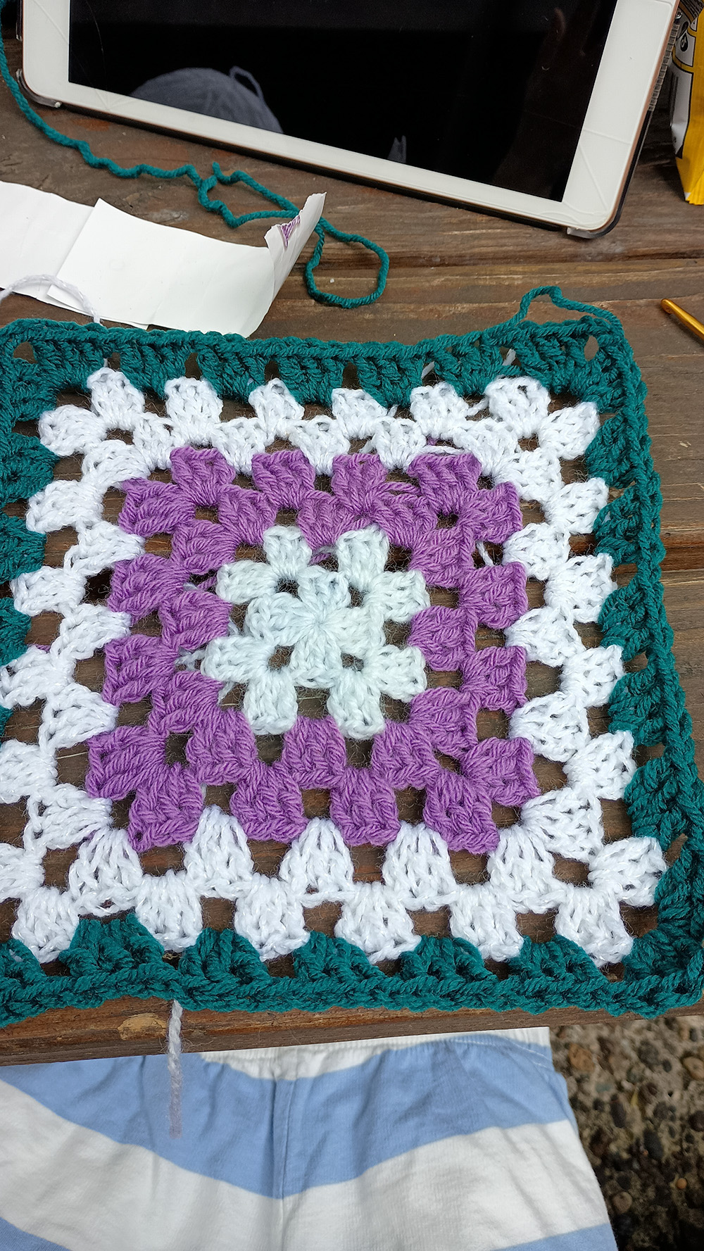 Crochet Granny Square A top-down view of a colorful handmade crochet granny square with a light blue center, purple and white middle rounds, and a dark teal border resting on a wooden surface.