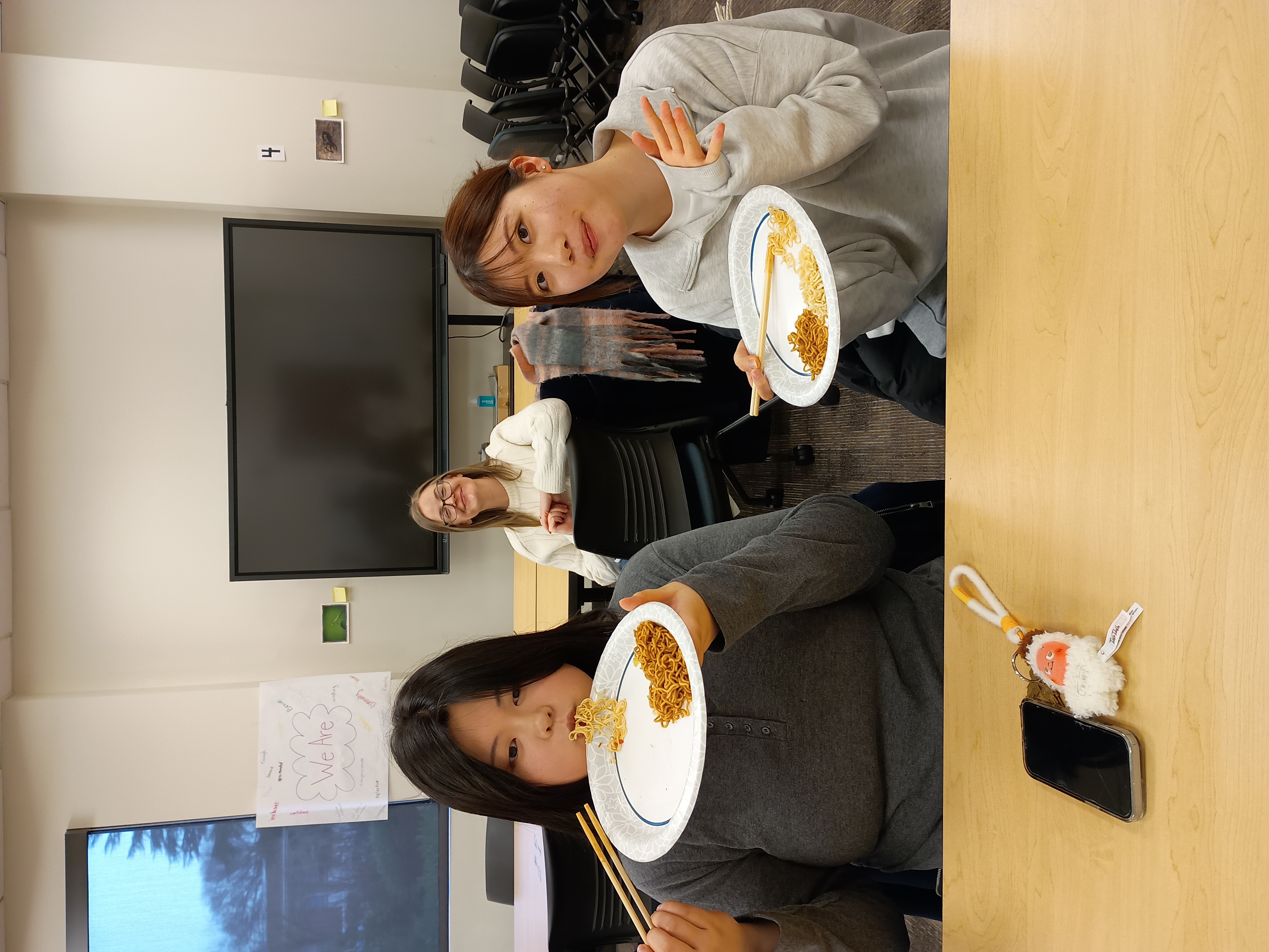 3 International students sitting in an on campus classroom and showing off their ramen dishes and smiling