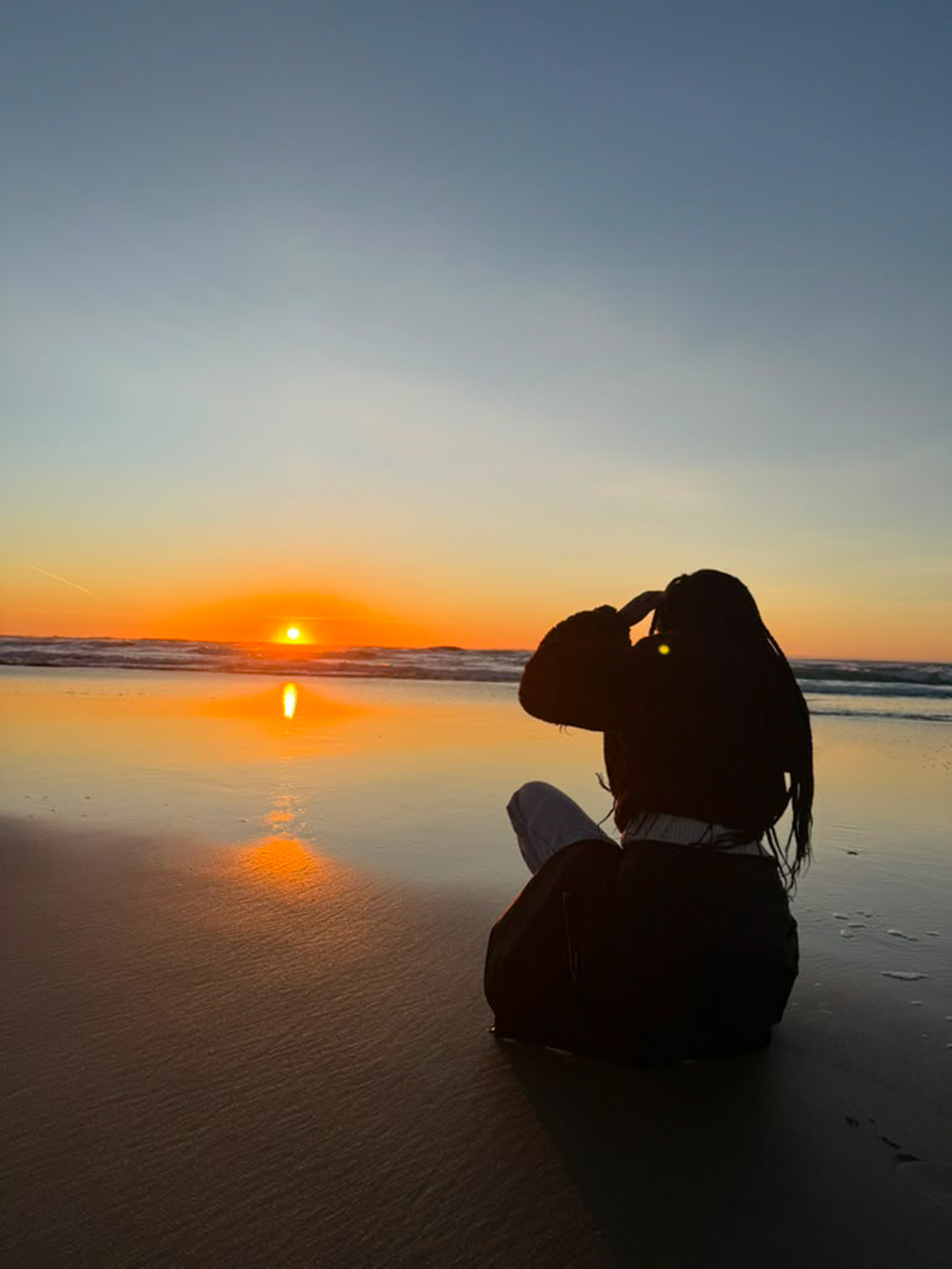 Jane ビーチで Jane 砂浜であぐらをかき、海に沈む鮮やかな夕日を眺める彼女の姿が、シルエットとして浮かび上がっている。