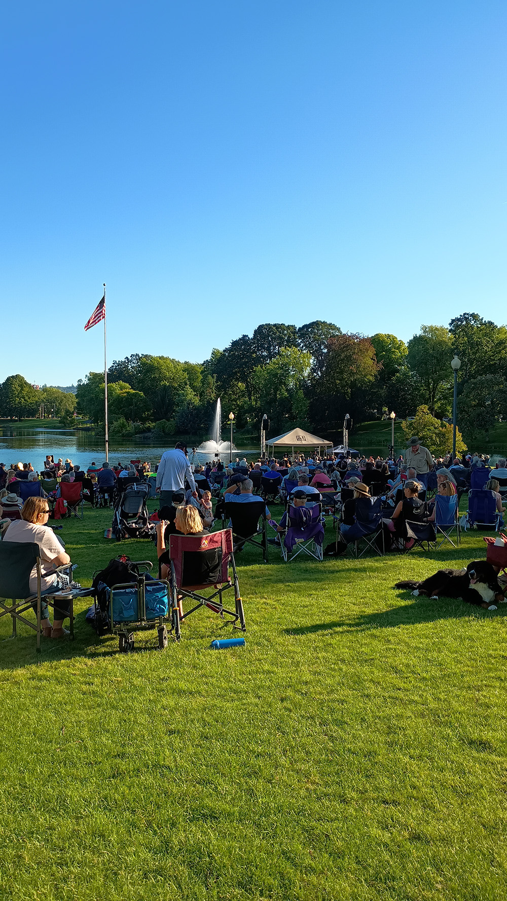 Concert at the Lake A large crowd of people sits in lawn chairs on a grassy hillside, watching an outdoor event near a lake with a fountain under a clear blue sky.