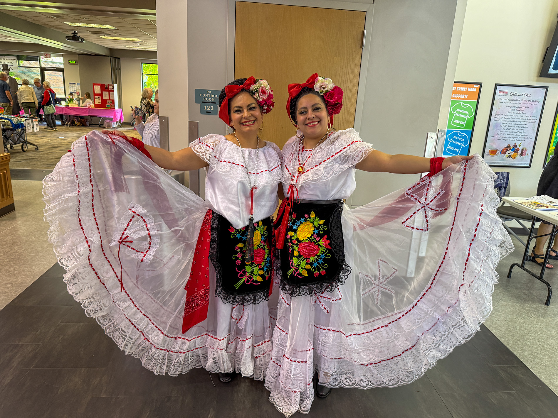 two performers wearing jarocho style dresses from Veracruz, Mexico, characterized by white lace-trimmed tiered skirts, embroidered black aprons, and floral headpieces.