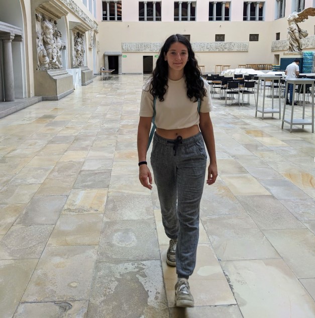 Studen ambassador, Fernanda, profile image walking down a stone pathway with tan and white buildings behind her wearing grey pants and white shirt