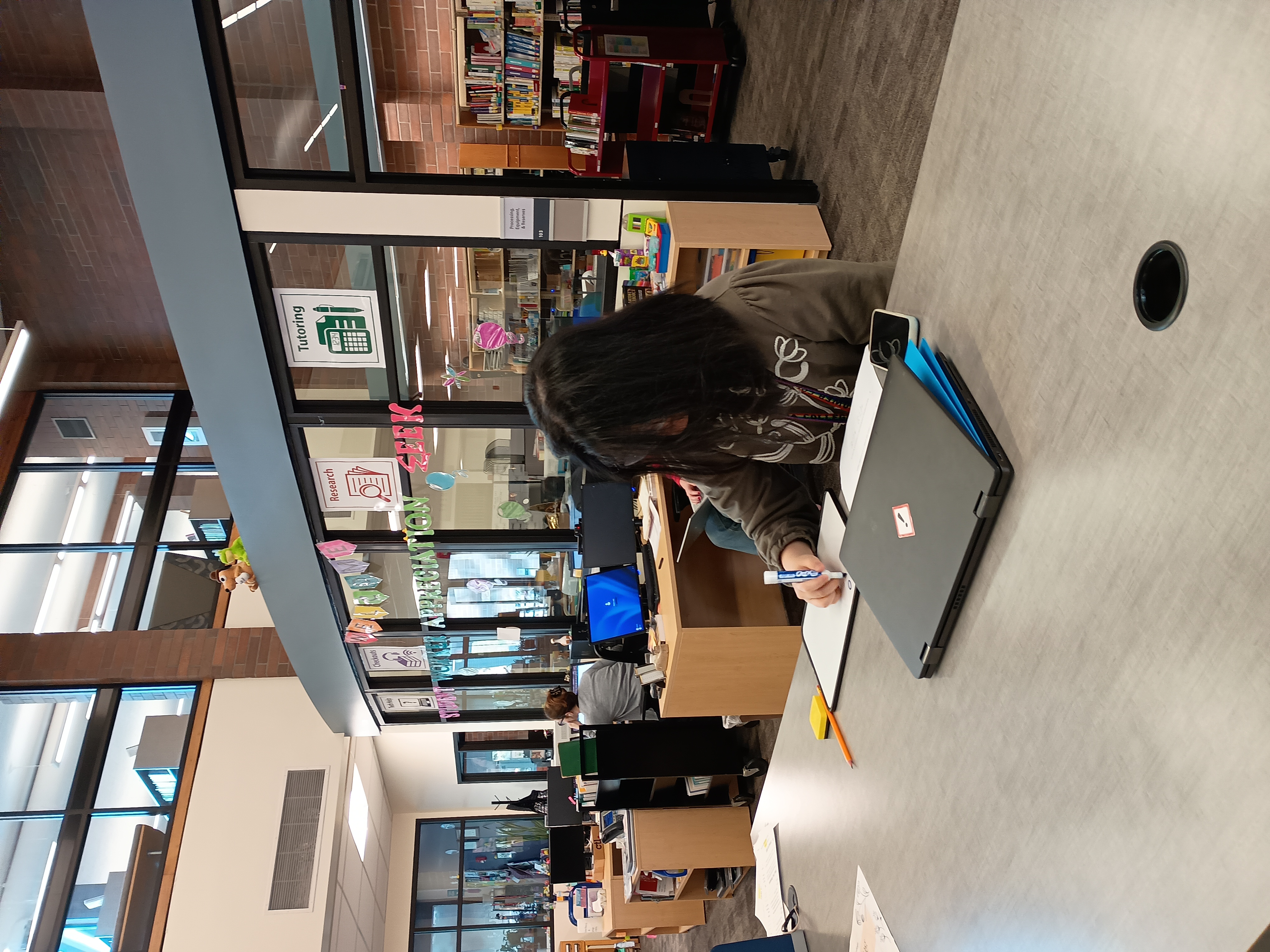 student worker in the Lower Columbia Library Learning commons with books and shelves in the backfround as the student worker looks down and is working on paperwork as a student tutor