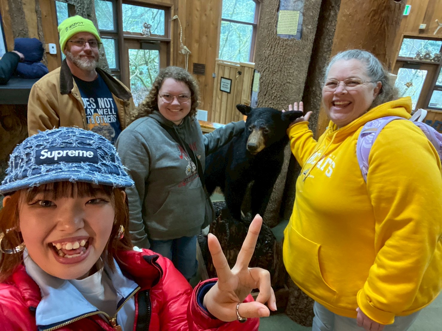 International student taking a selfie holding up a piece sign and smiling with two females and a male (host family) in the background showcasing a taxidermy black bear.