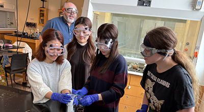4 international students in a science lab with blue rubber gloves and goggles on running and experiemnt in a beaker with the instructor walking by in the background