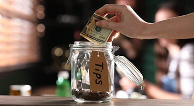 Tip jar on a storefront counter and someones hand holding money putting it in the jar with out of focus baristas in the background