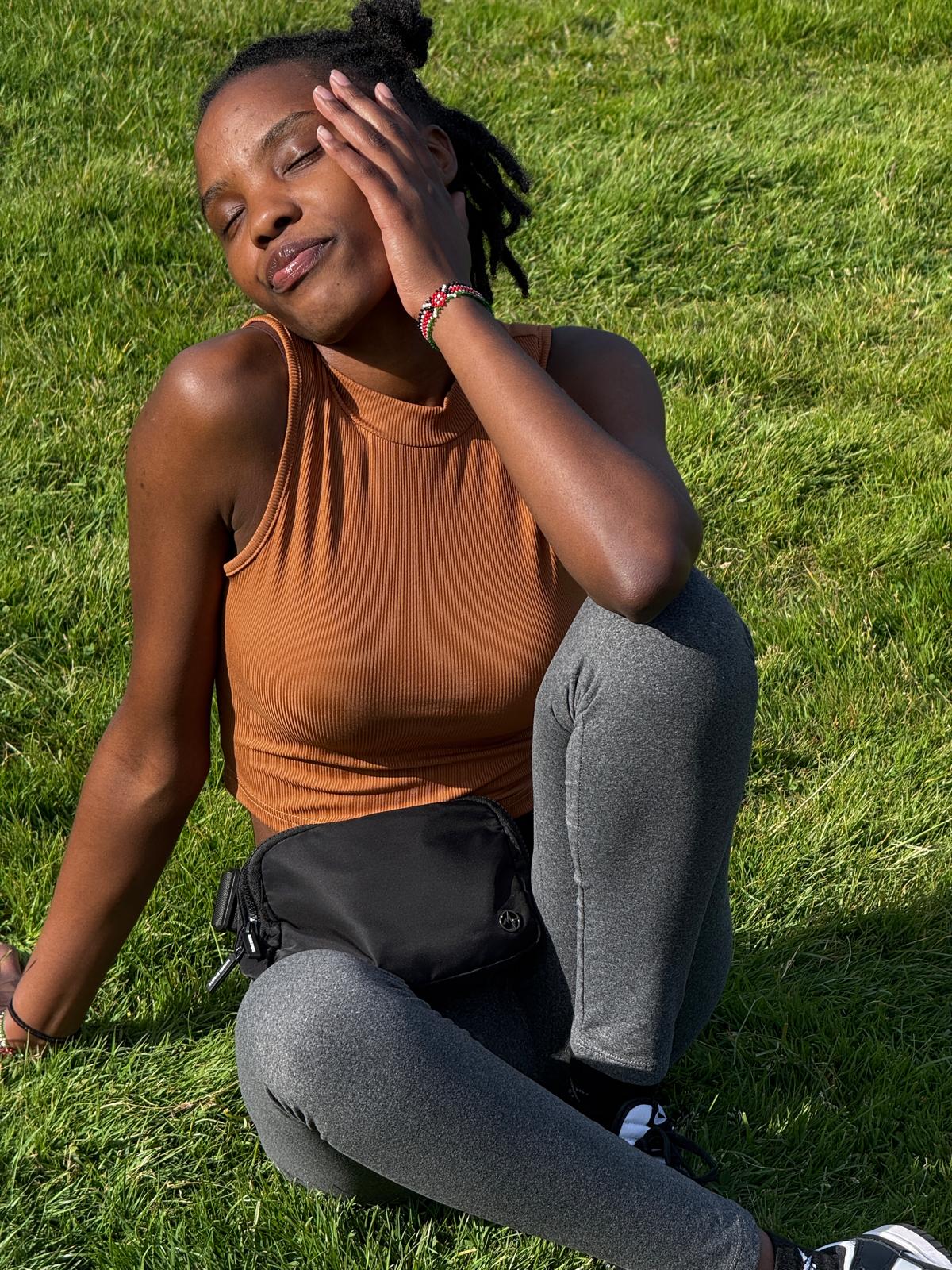 Jane, International Student from Kenya sitting on the grass on a sunny day with her eyes closed and smiling wearing an orange tanktop and dakr jeans