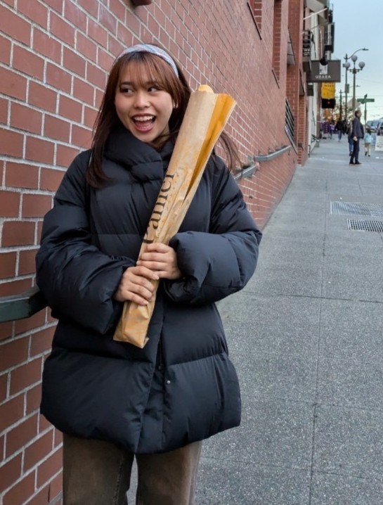 International Student from Lower Columbia College Rinna. Rinna has shoulder-length auburn hair wearing a black puffer jacket, holding a baguette wrapped in paper. She is smiling and looking to the right, standing on a sidewalk next to a brick building. In the background, other pedestrians are walking on the sidewalk.