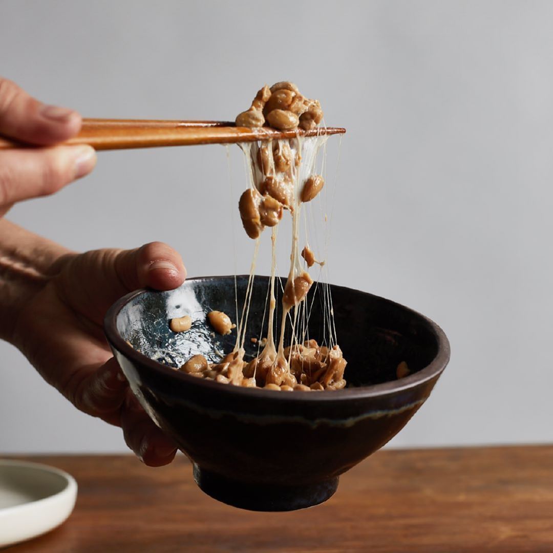A person is lifting natto, a sticky fermented soybean dish, from a dark ceramic bowl using wooden chopsticks, creating long, stringy filaments. The bowl is held over a wooden table.