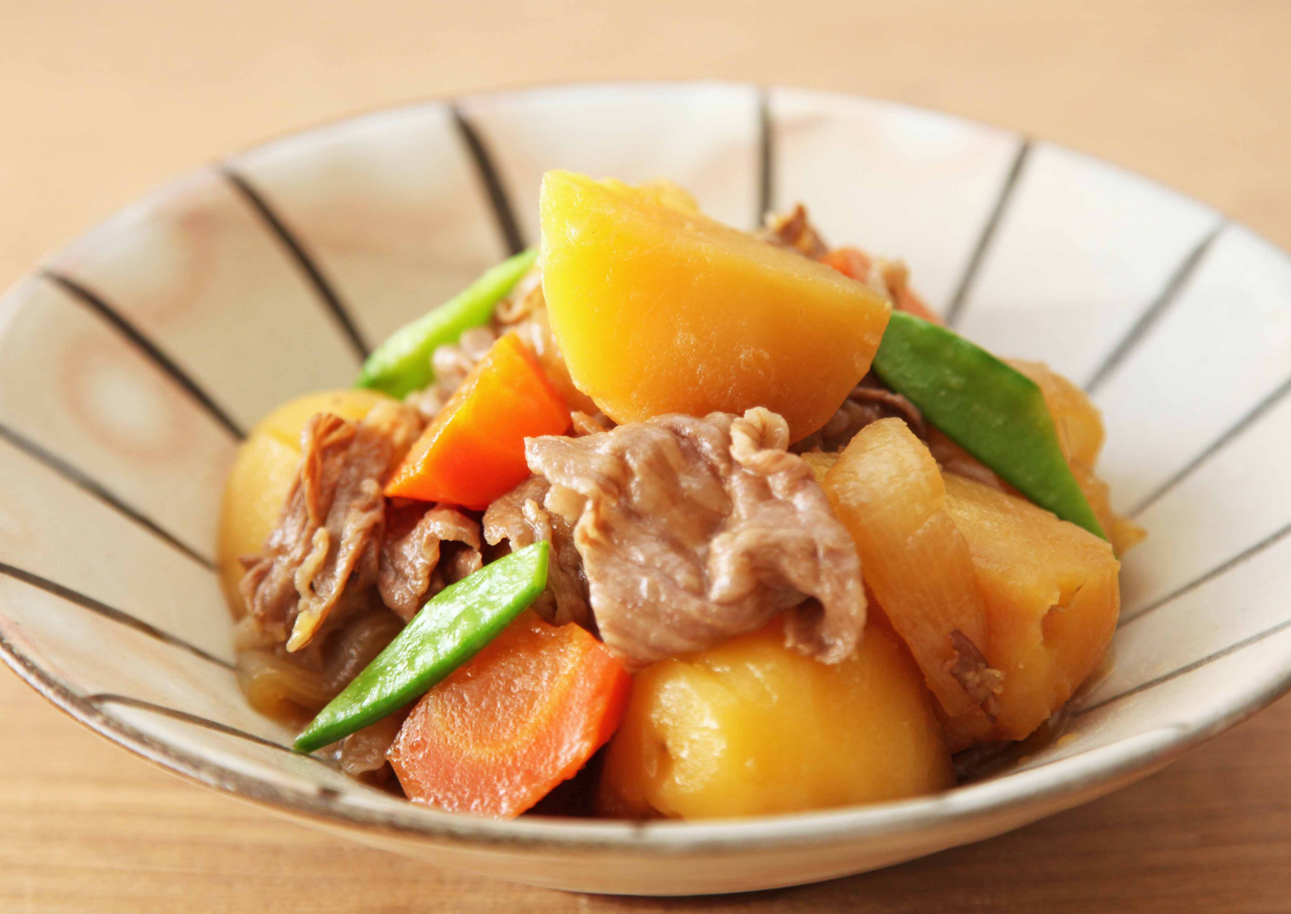 A close-up photograph of a bowl of Japanese Nikujaga stew, containing chunks of potato, sliced beef, carrots, and snow peas, served in a ceramic bowl with brown stripes on a wooden table.