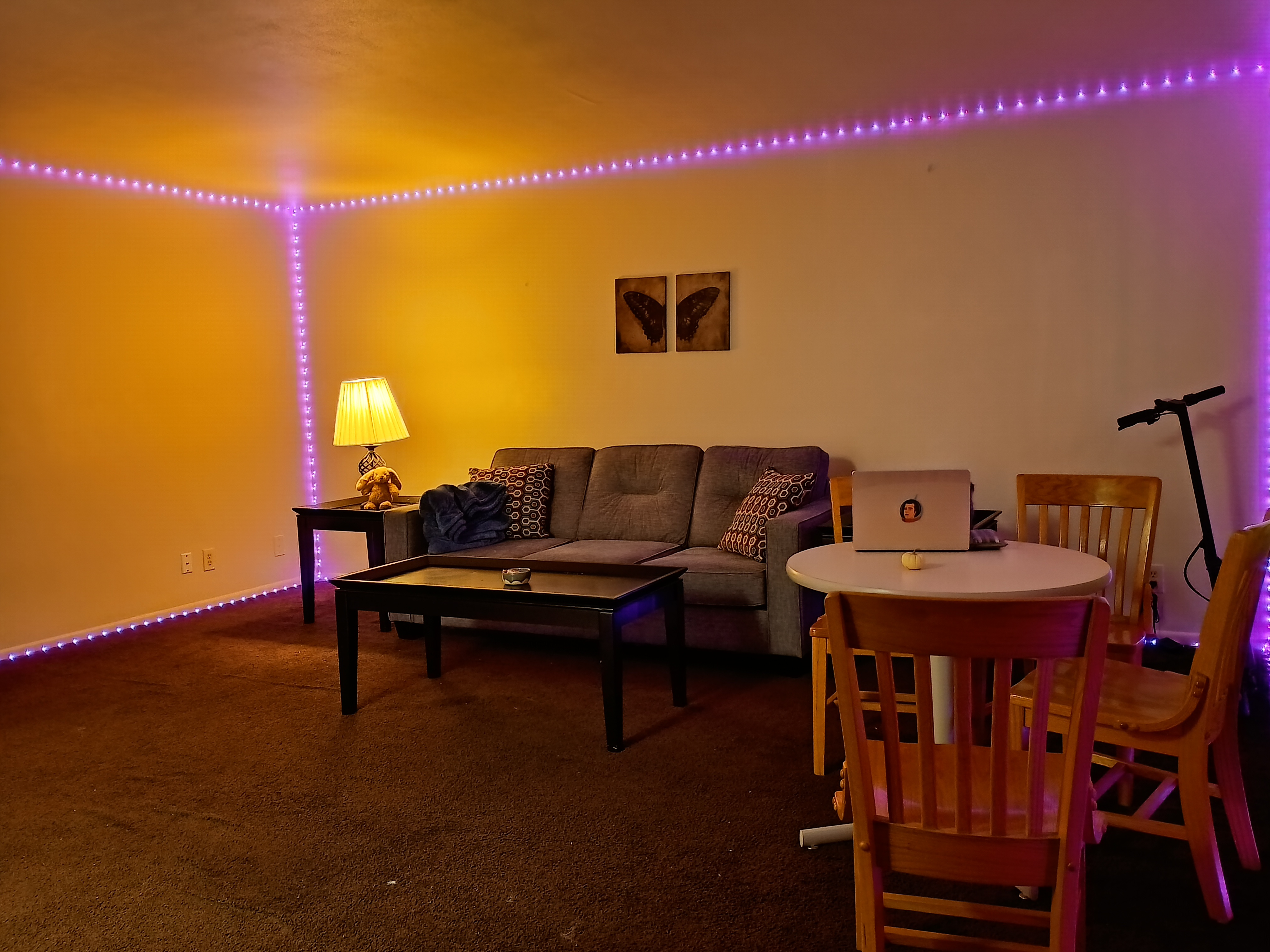 International Student housing living room with low lighting from a lamp in the corner. The room has a lamp, couch, small round table with four chairs, an electic scooter leaning against the wall and rope lights ran along the ceiling.