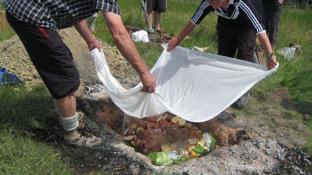 Two people covering Hangi in a hole in the ground with a white sheet.