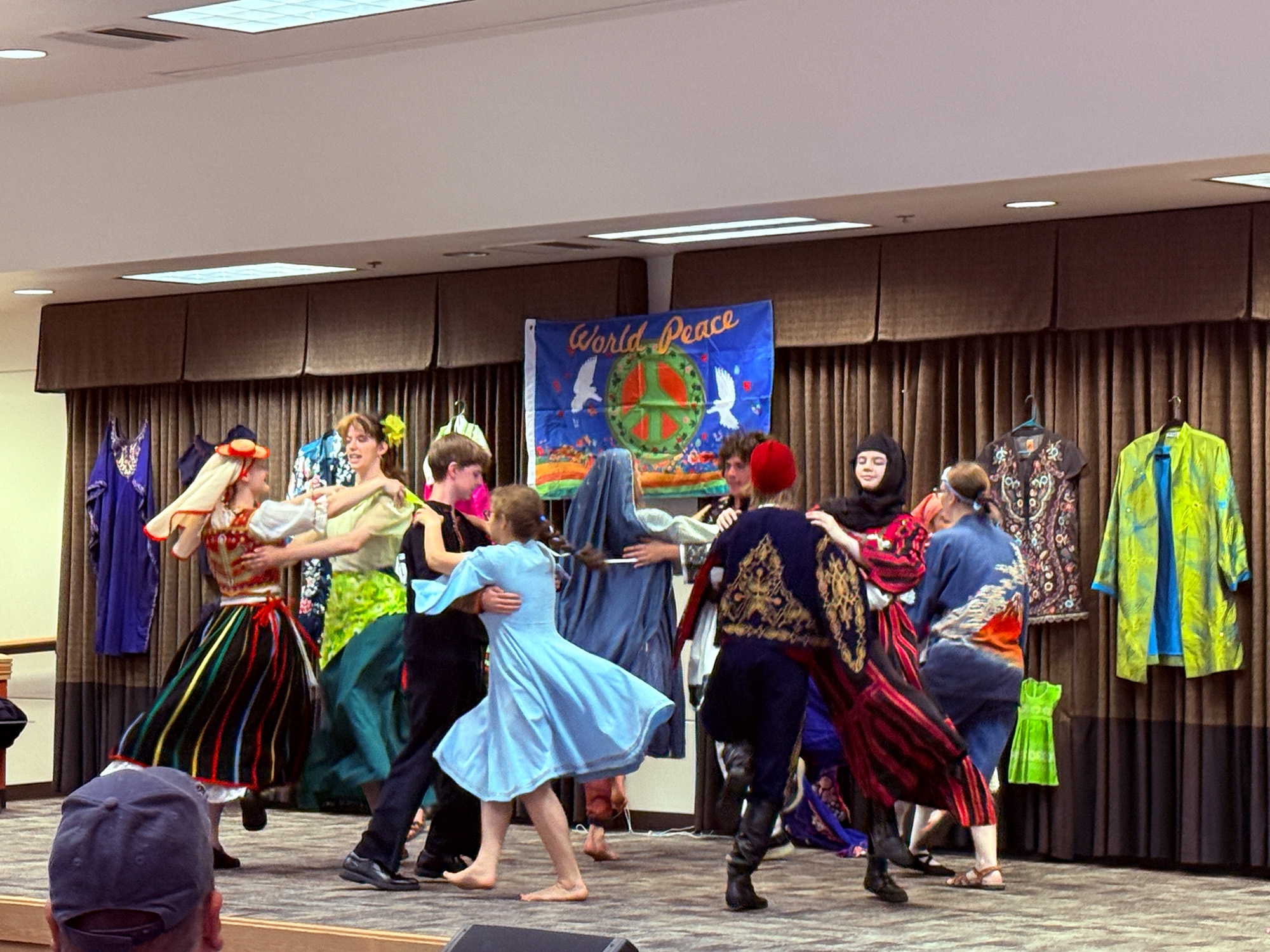 a group of performers from the Sussefusse International Folk Dance Troupe in diverse cultural attire, engaging in a traditional folk dance on a stage with a "World Peace" banner in the background.