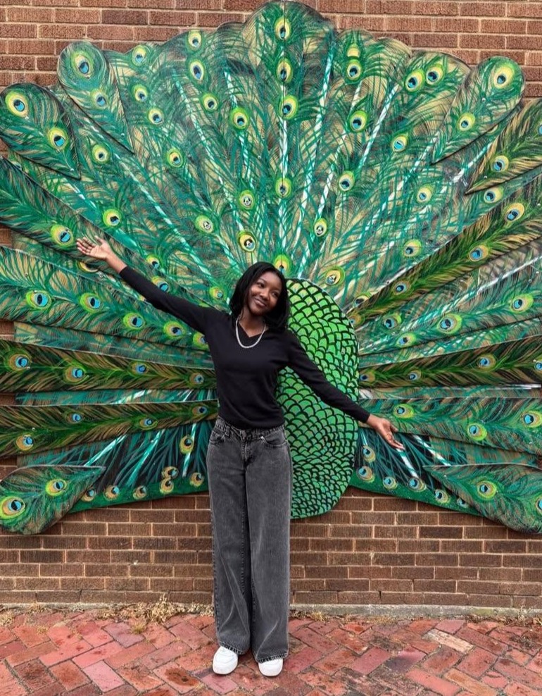 picture of student standing in front of peacock feathers displayed on a brick wall