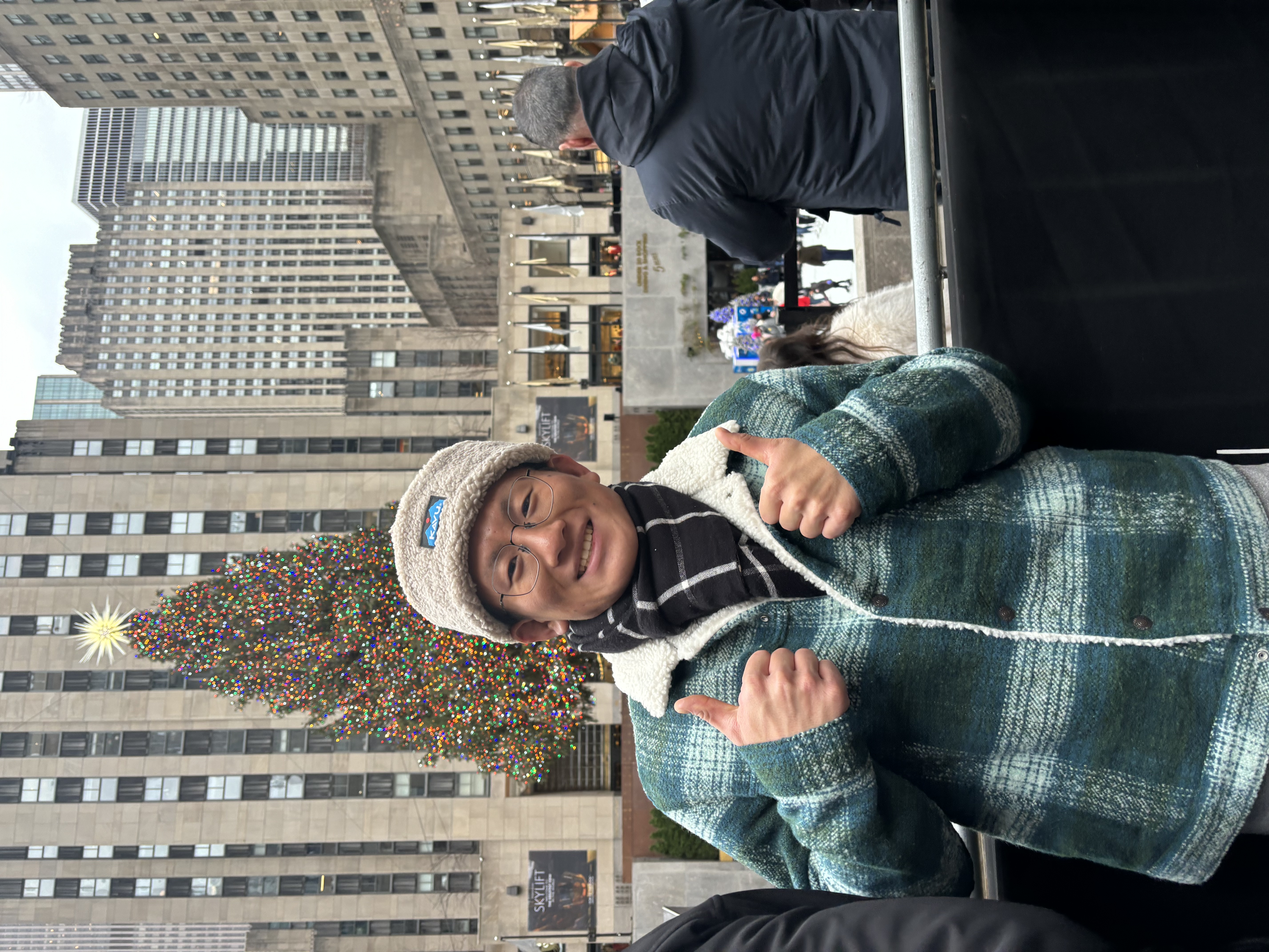 International student Jingru posing for an image wearing a plaid jacket and scarf and gray cap, smiling and giving a double thumbs up.