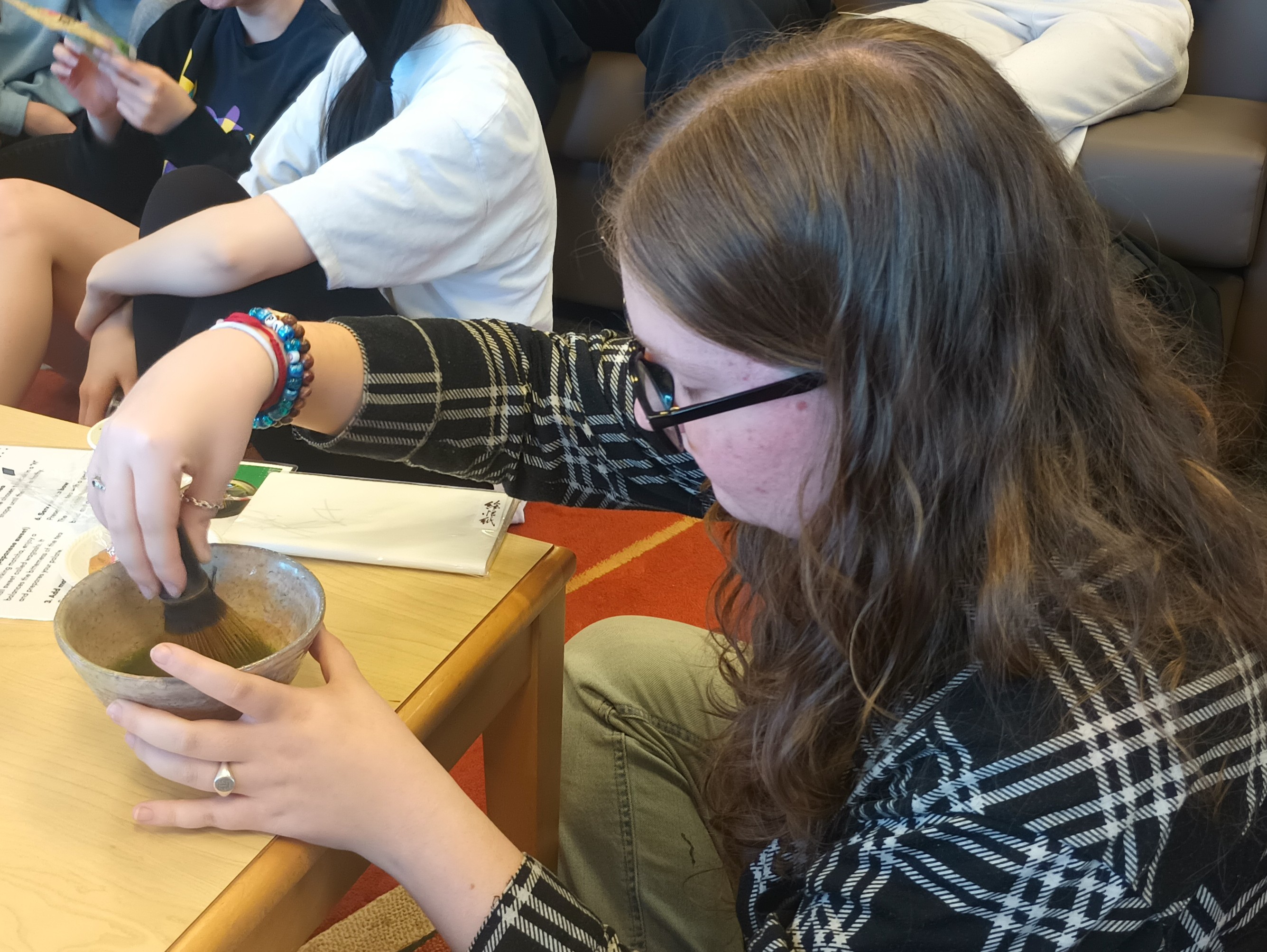 female college student trying to make her own ceremonial macha. Sitting at a coffee table and other students in the background