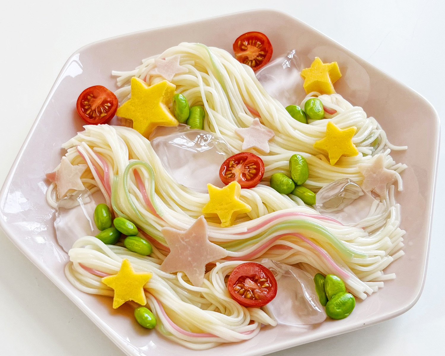 A ceramic bowl filled with somen, halved cherry tomatoes, edamame, slices of pork in the shape of stars, and carrot slices in the shape of stars.