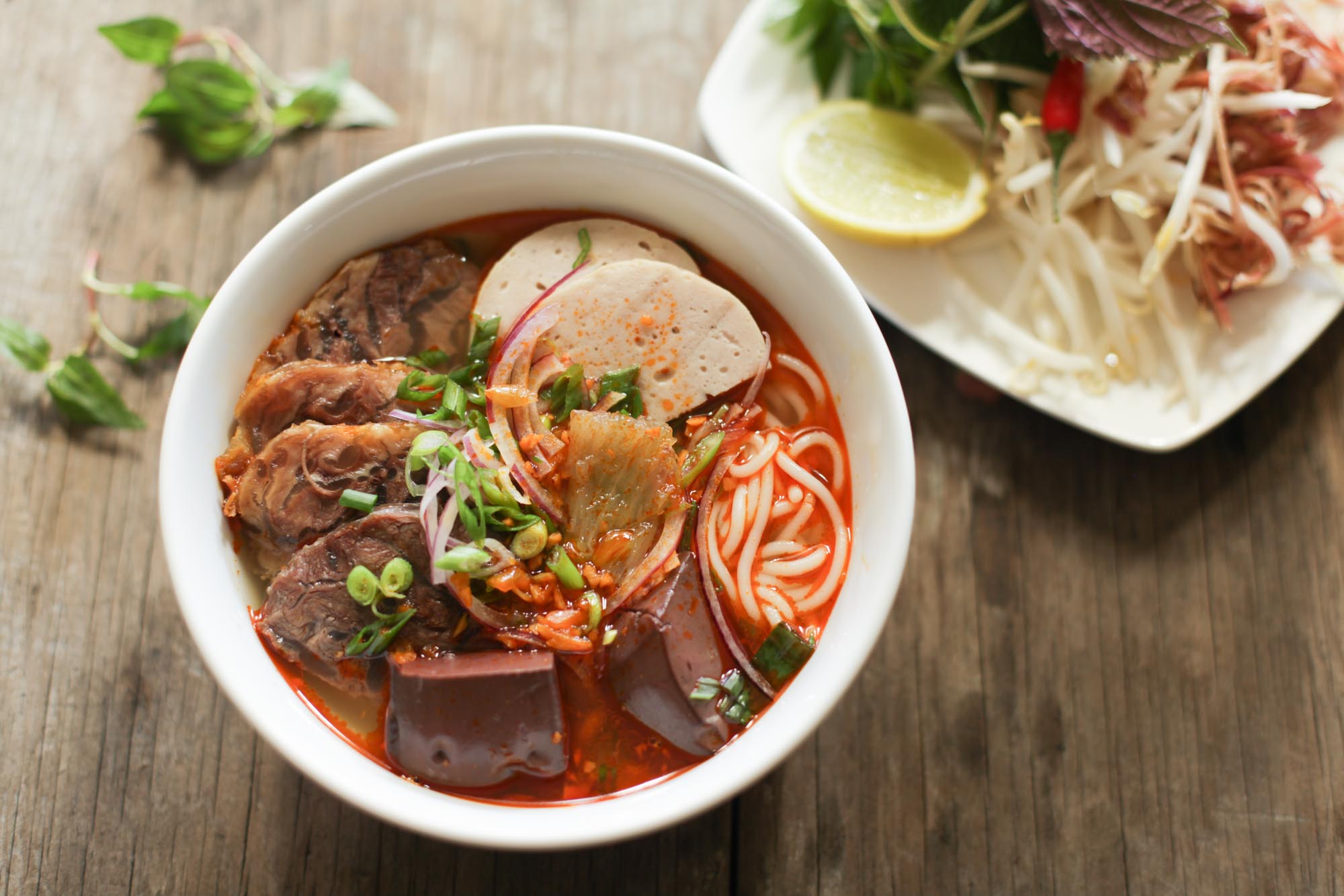 A bowl of Vietnamese Bun Bo Hue, a spicy beef noodle soup, is presented in a white ceramic bowl on a rustic wooden table. The dish features thick white rice noodles in a rich, reddish-brown broth, topped with slices of braised beef shank, pork sausage (chả lụa), cubes of congealed pig's blood, sliced red onions, scallions, and cilantro. A side plate holds bean sprouts, lime wedges, fresh herbs, and shredded banana blossom.