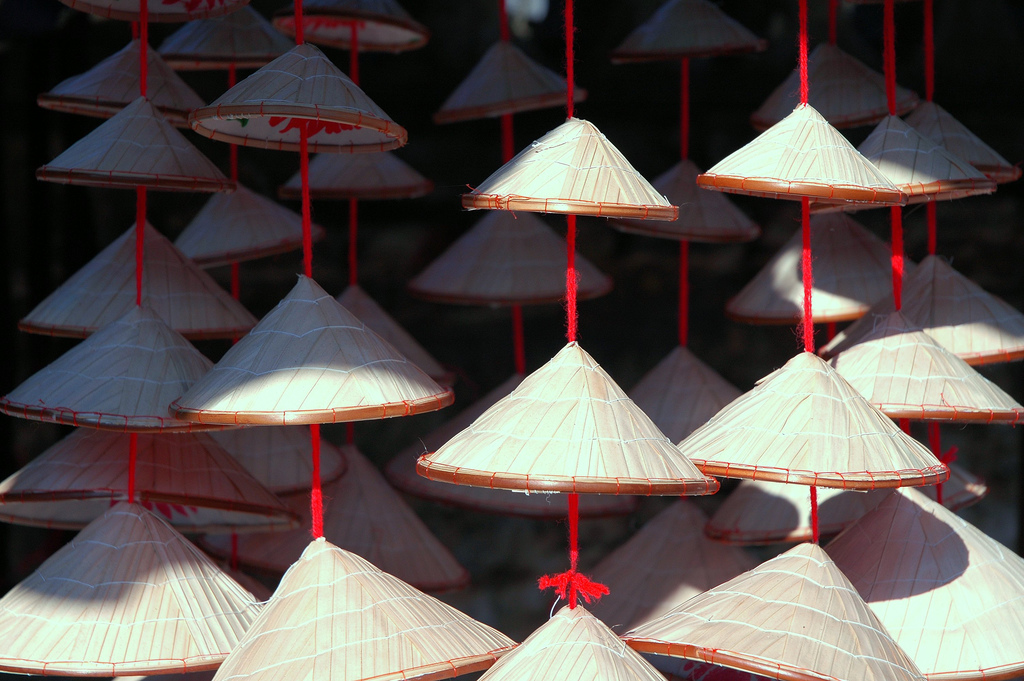A photograph captures numerous light-colored, conical straw hats, known as nón lá, hanging by red strings in rows. The hats are woven from dried leaves and have a wide, circular brim with a pointed top. Some hats are brightly lit, while others are in shadow, against a dark, out-of-focus background.