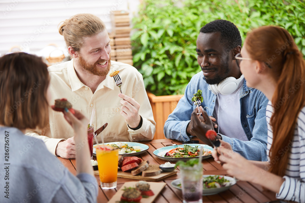 4 people, 2 males and 2 females, sitting outside at a wooden table eating salads and looking at one another smiling and laughing
