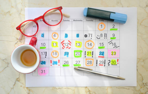 Printed calendar with highlights and notes. Empty cofee cup sitting next to it with pens and red glasses.