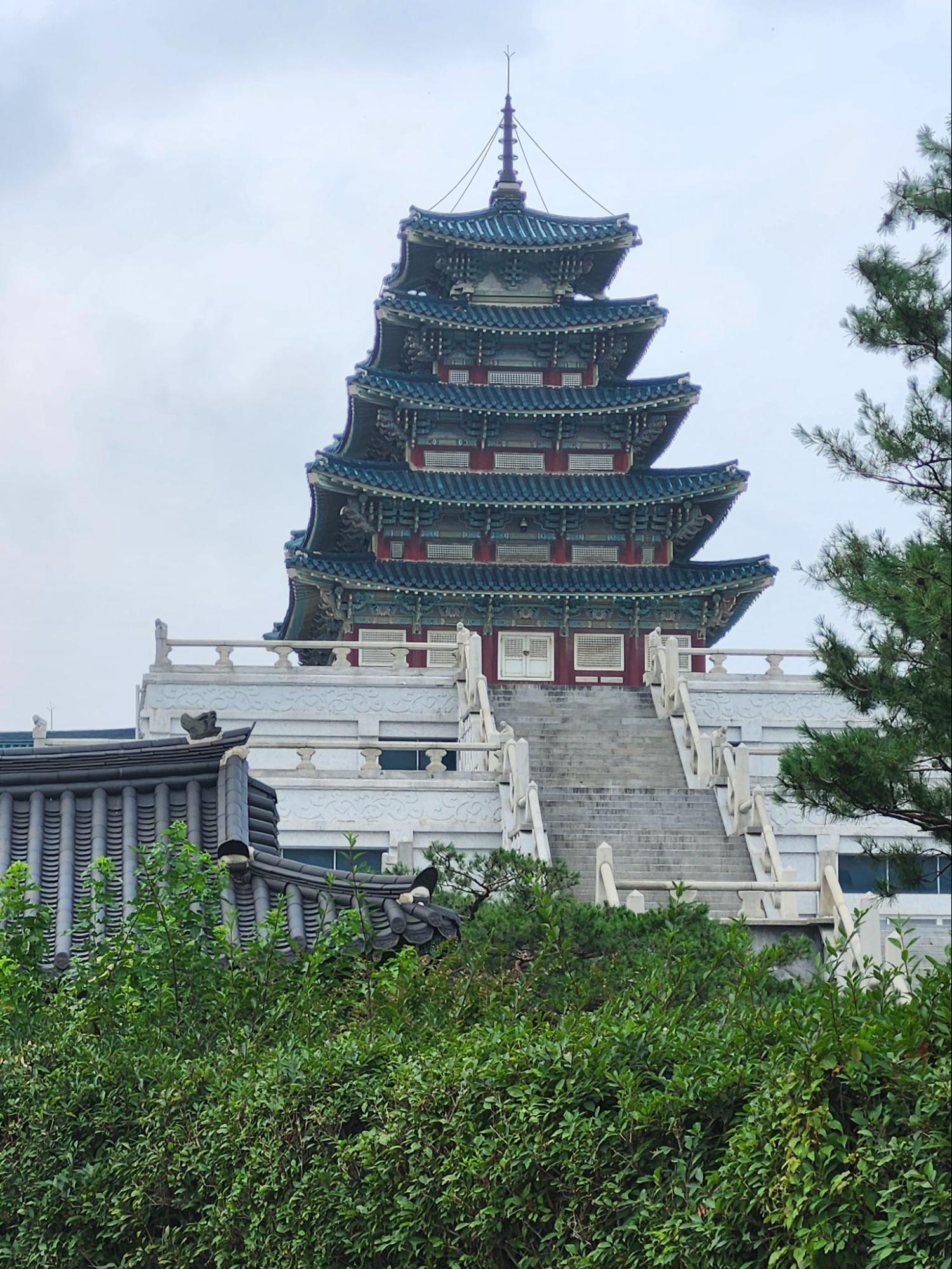 A vertical shot of the National Folk Museum of Korea pagoda in Seoul. The multi-tiered structure features vibrant blue-green tiled roofs and red accents, sitting atop a white stone base with a grand central staircase. Lush green bushes and the dark-tiled roof of a traditional gate are visible in the foreground under an overcast sky.