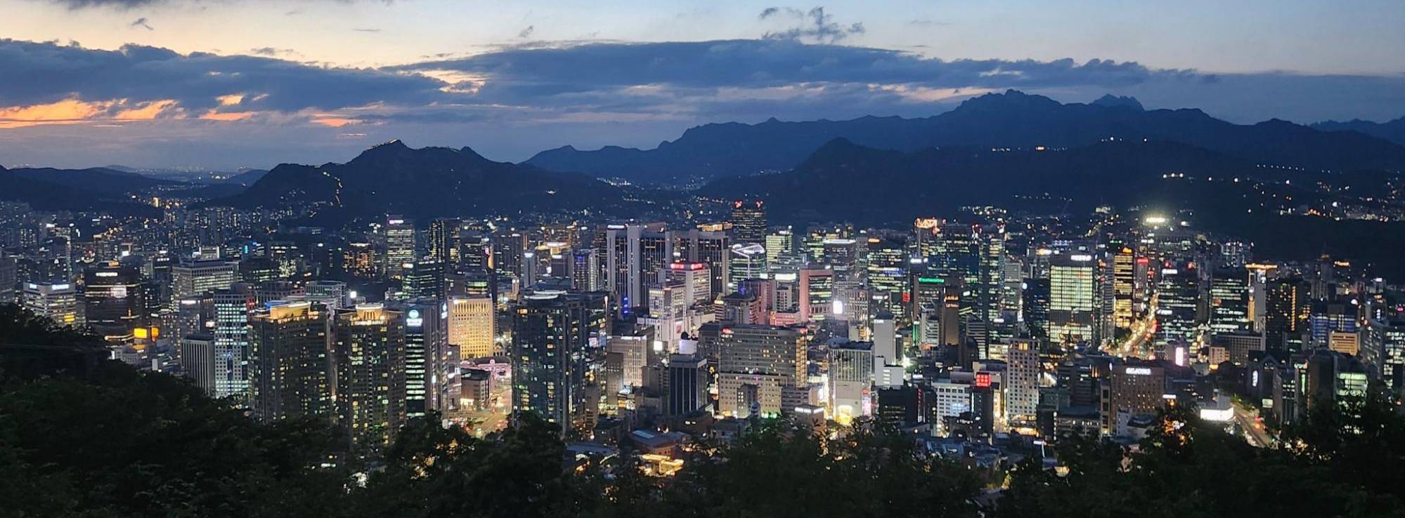 A wide-angle, high-elevation view of Seoul at twilight. Thousands of lights from city skyscrapers and office buildings glow in the valley below, framed by the dark, silhouettes of jagged mountains in the background. The sky is a deep blue with streaks of orange from the setting sun and scattered dark clouds.