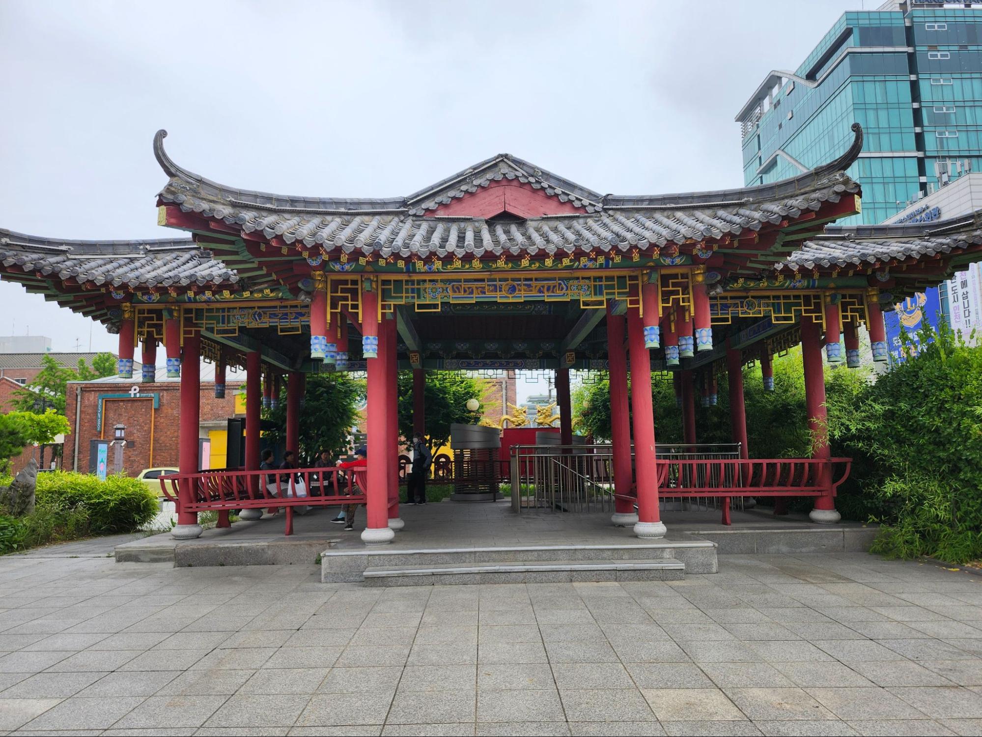A wide shot of a traditional Chinese-style pavilion located in Incheon Chinatown. The structure features a gray-tiled roof with upturned eaves, bright red pillars, and intricate yellow and blue decorative carvings. In the background, a modern glass office building creates a sharp contrast between traditional and contemporary architecture.