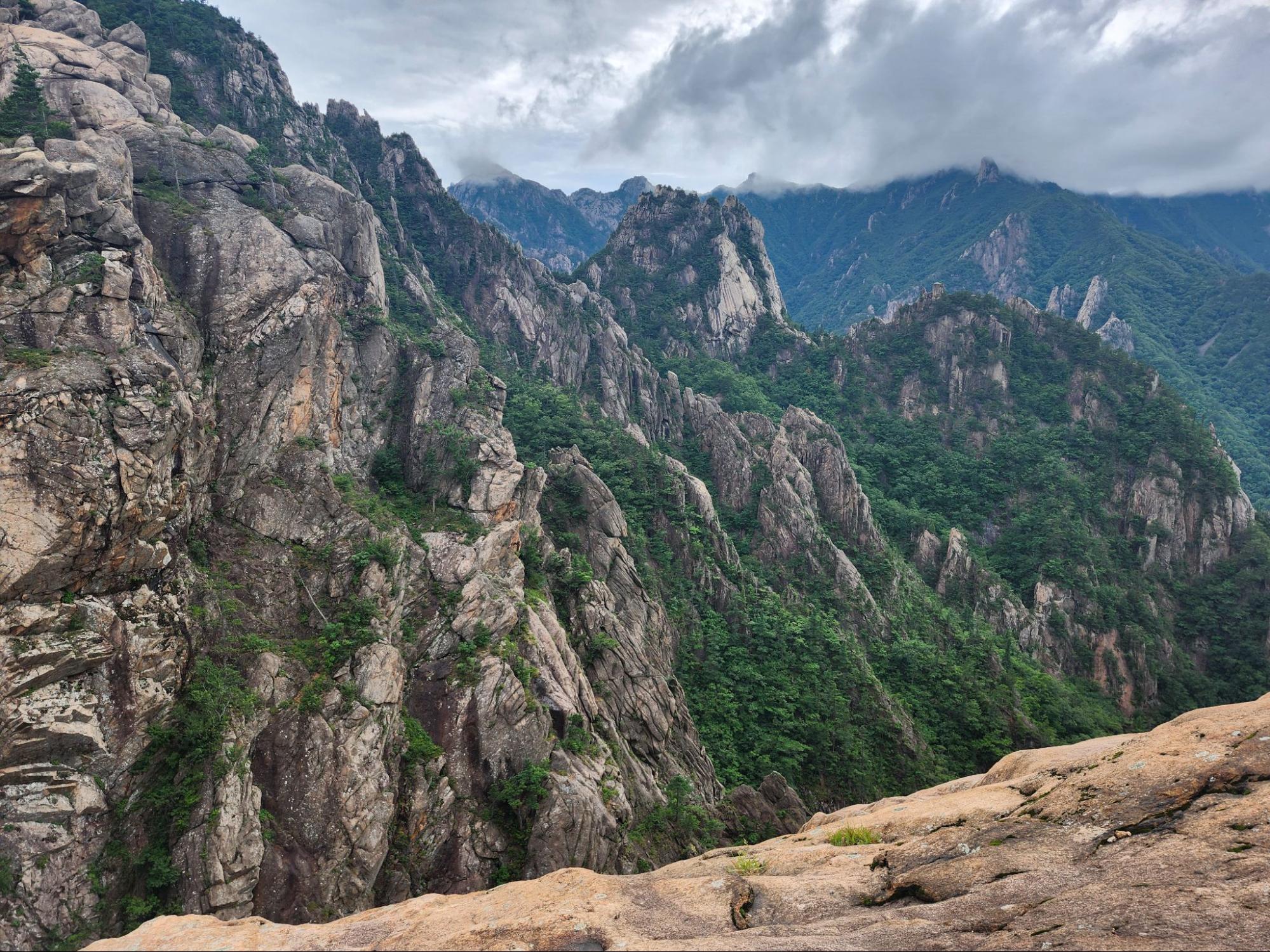A breathtaking landscape view of the rocky peaks of Seoraksan National Park. The rugged, light-gray granite cliffs are interspersed with patches of deep green forest. Thick, low-hanging clouds partially obscure the distant mountain ridges, creating a moody and dramatic atmosphere.