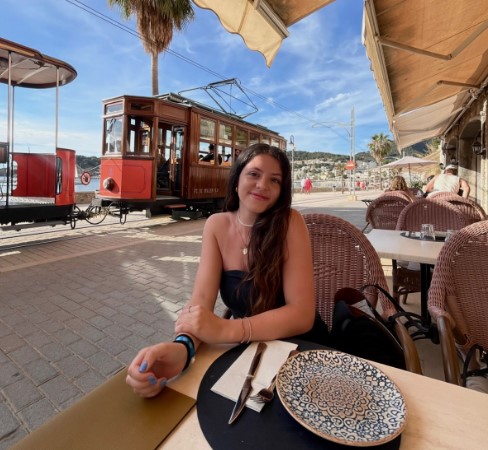 LCC Student ambassador, Lisa, posing at a outdoor dining table with a trolly and beach in the background