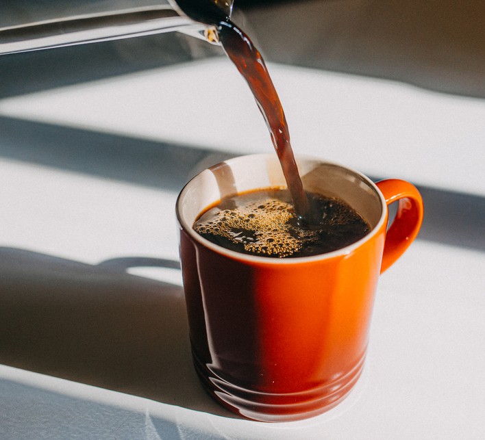 An orange and brown ceramic cup sitting on a table with hot dark coffee being poored into it