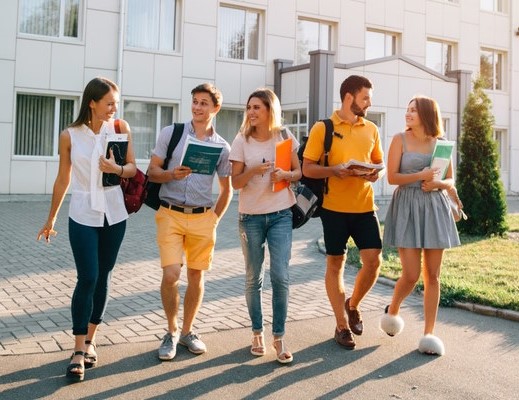 College aged students walking together on a campus while holding books on a sunny day.