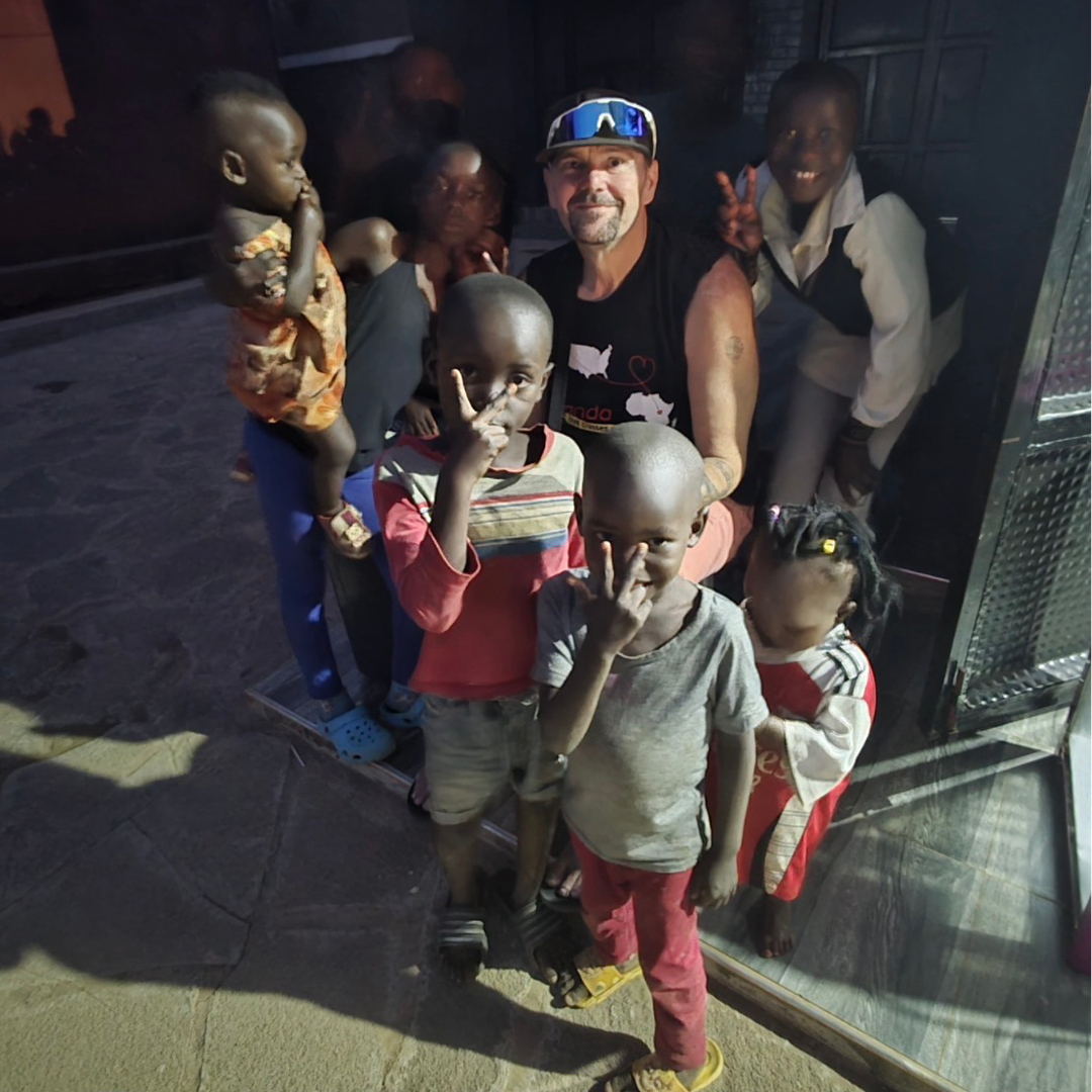 LCC Adult Basic Education graduate Craig Millar, wearing a visor and tank top, smiles surrounded by several young children making peace signs while engaging in non-profit community work in Uganda.