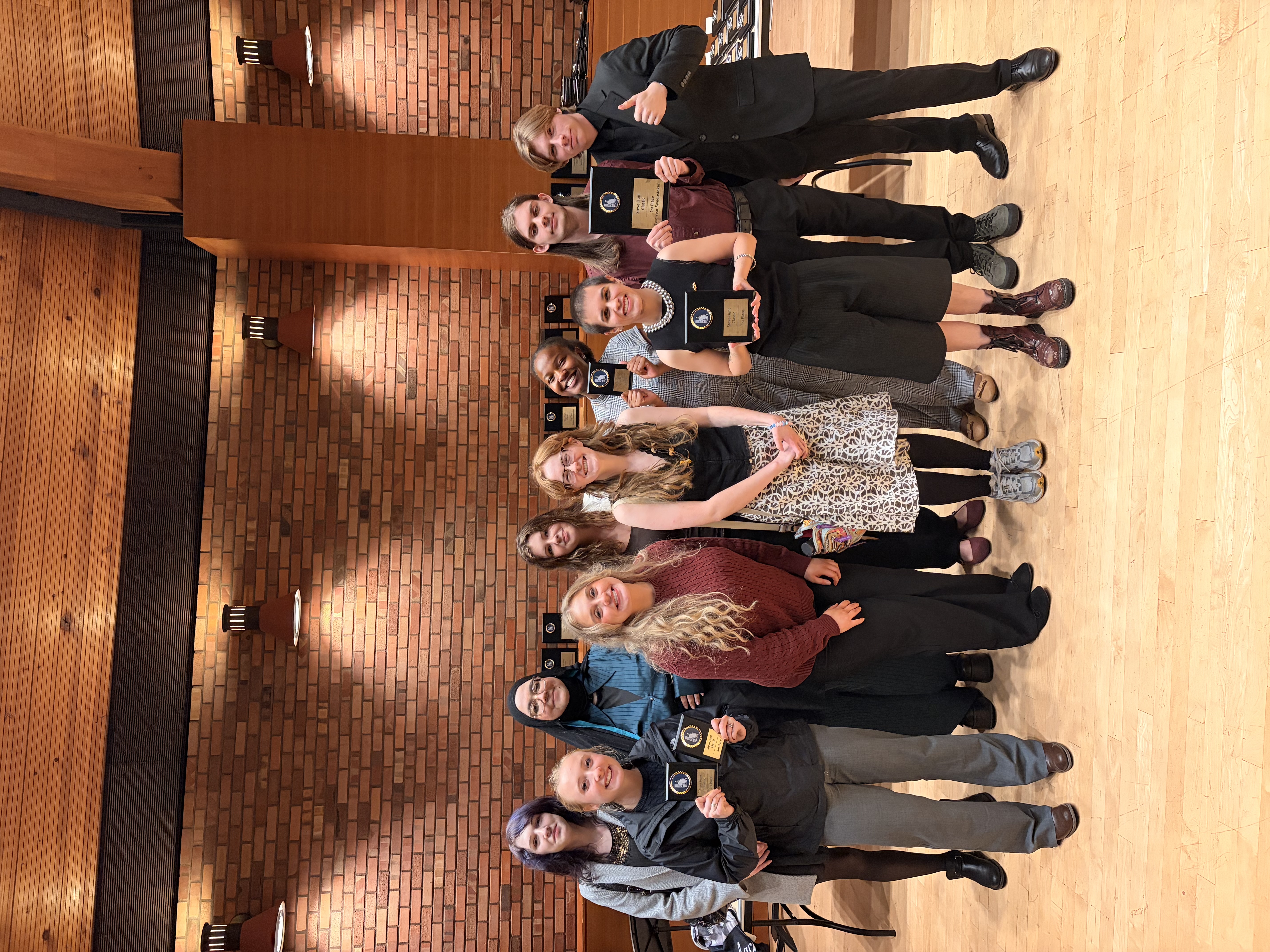 ten diverse young adults posing happily in an indoor ceremonial space, likely an auditorium or hall. Four individuals in the front-center-left and front-center-right are holding black plaques with gold inscriptions, which they proudly display towards the camera.They are standing on a light-colored wood floor in front of a textured red brick wall with two prominent wall sconces. Wooden ceiling panels are visible above, along with more brickwork.
