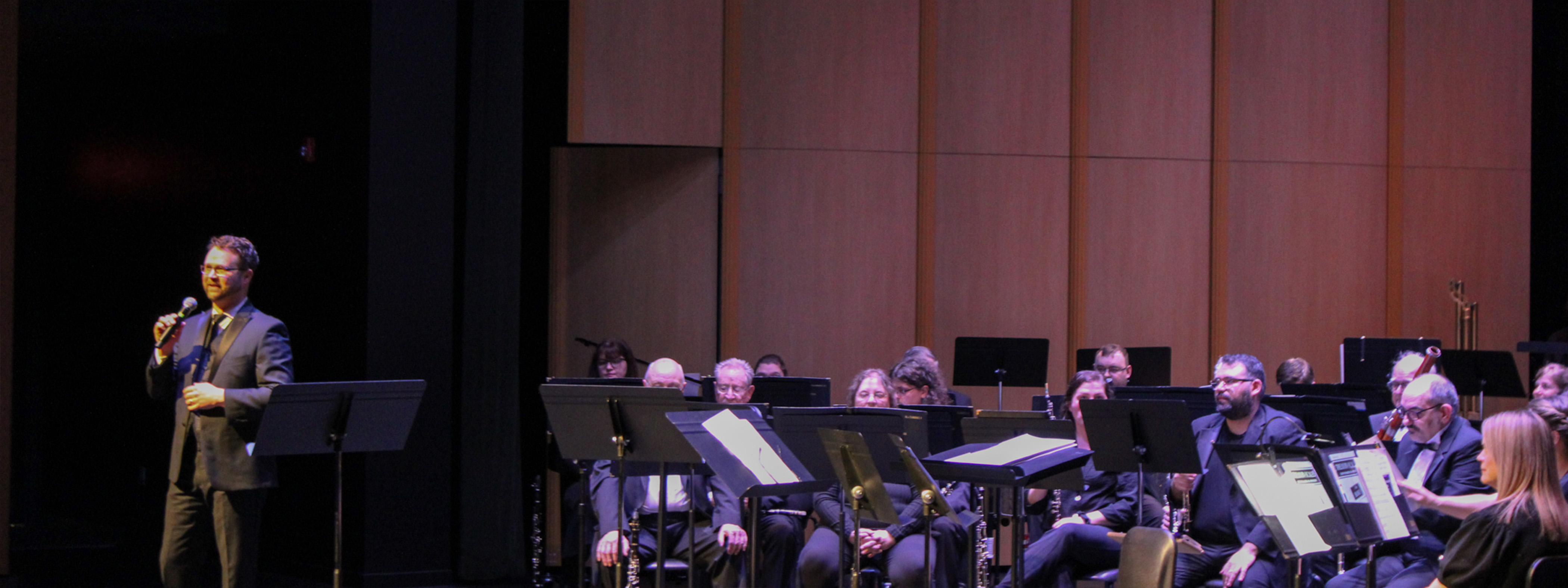 Members of the symphonic band waiting before a performance as the director stands at the front of the stage giving the pre-show speech.